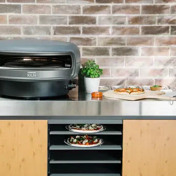 Everdure Kiln S vs Everdure Kiln R graphite color oven on stand in front of brick wall