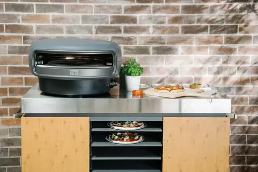 Everdure Kiln S vs Everdure Kiln R graphite color oven on stand in front of brick wall